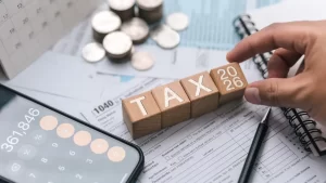 Wooden blocks spelling TAX 2026 on a tax form with calculator and coins, representing NRI property TDS compliance changes in 2026.