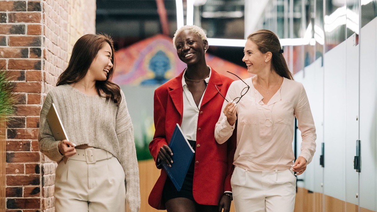 HPI Visa FAQs Three young professional women walking and talking in a modern office, illustrating questions about the UK High Potential Individual visa
